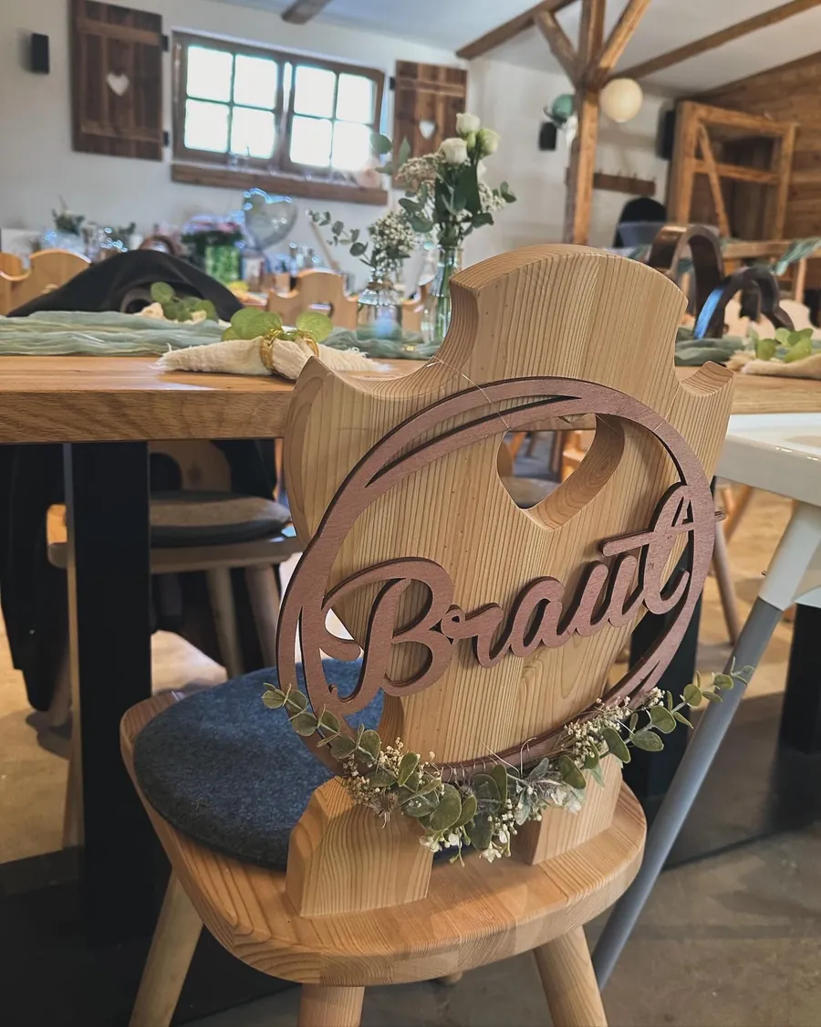 Alm-Stuhl mit Herzausschnitt und Holzschild „Braut“ mit Eukalyptuskranz, dahinter die festlich gedeckte Hochzeitstafel